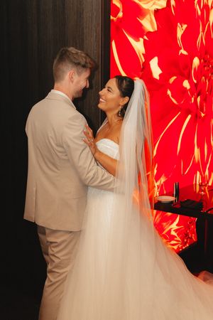 Bride in white strapless gown and veil smiling at groom in beige suit before red floral backdrop.