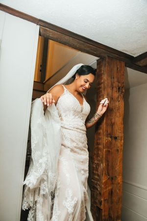 Smiling bride in a lace wedding gown and veil standing near rustic wooden beams indoors.