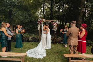 Two brides sharing a first kiss at an outdoor woodland wedding ceremony with a floral arch.