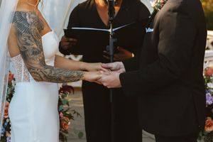 Bride with tattooed arms exchanging vows and rings with groom before officiant at outdoor wedding ceremony.