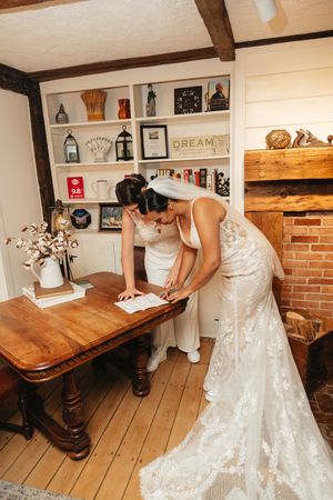 Bride in a lace gown and veil signing wedding documents at a wooden table in a rustic room.
