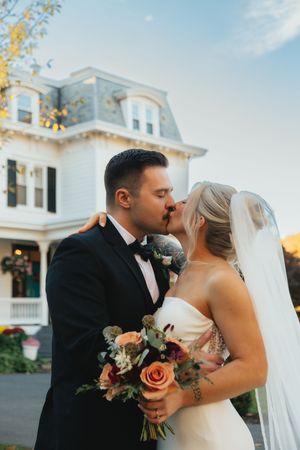 Bride and groom sharing a romantic kiss outside a Victorian mansion, bride holding a lush autumn bouquet.
