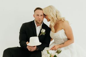 Bride and groom posing with a small white wedding cake, bride whispering in groom's ear during wedding portrait.