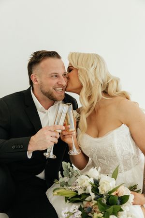 Smiling groom and blonde bride sharing a kiss while toasting with champagne glasses on their wedding day.