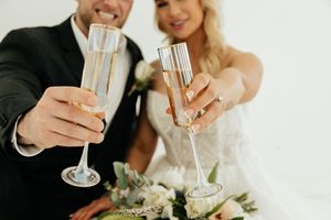 Close-up of bride and groom clinking elegant champagne flutes with wedding bouquet of white flowers nearby.