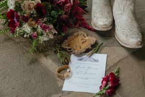 Wedding flat lay featuring a bouquet, white cowboy boots, rings, and elegant invitation on stone.