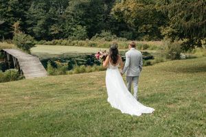 Bride and groom walking hand in hand across a lush green lawn toward a wooden bridge.