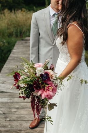 Bride holding a lush burgundy and pink bridal bouquet with ferns and greenery on a wooden bridge.