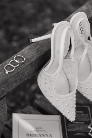 Black and white photo of pearl-studded ALDO bridal heels with wedding rings and invitation on wooden bench.