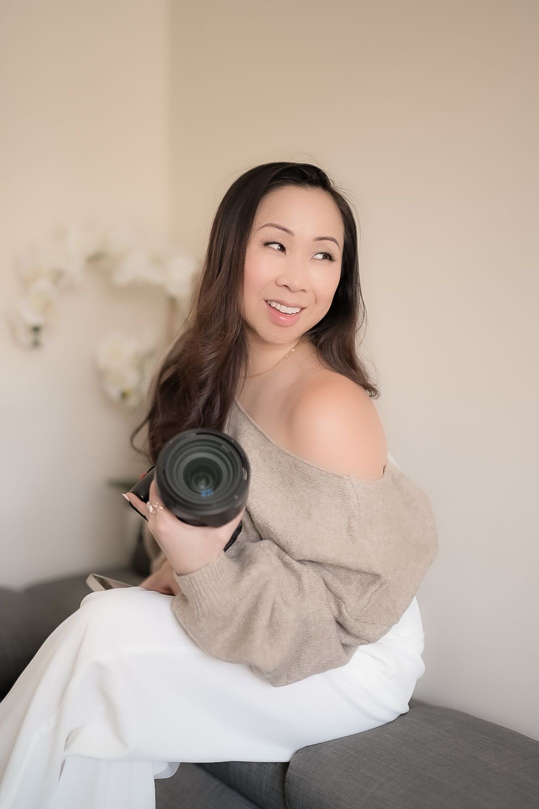 Professional photographer in cozy beige sweater holding camera lens while sitting on gray couch at home studio.