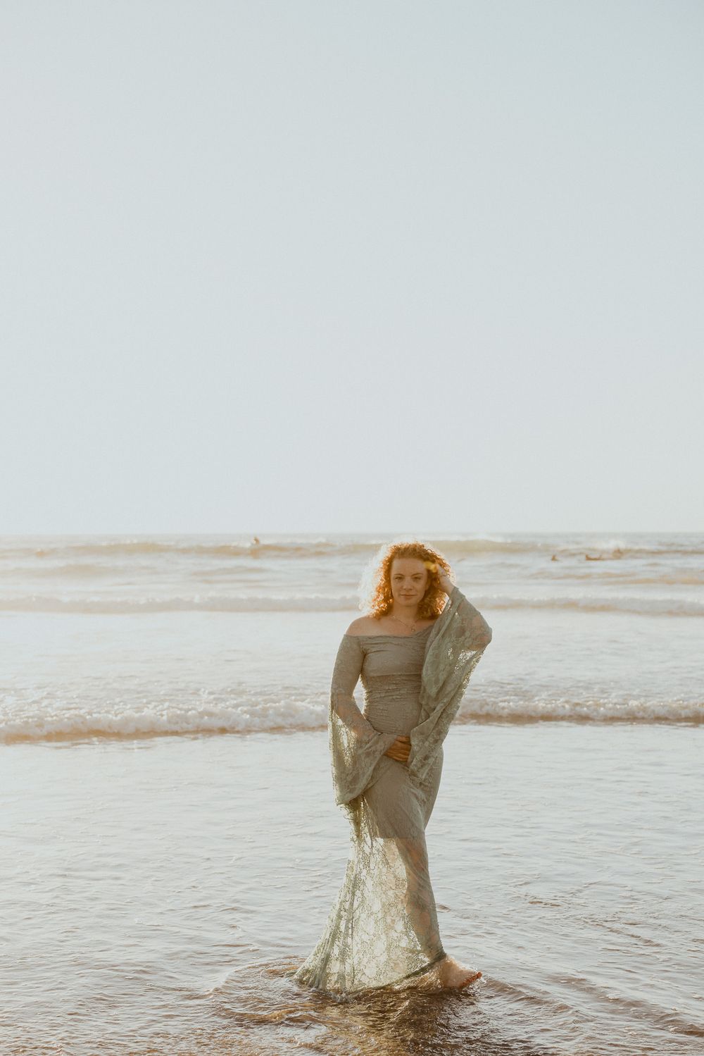 Person in a flowing dress standing at the ocean's edge during golden hour.