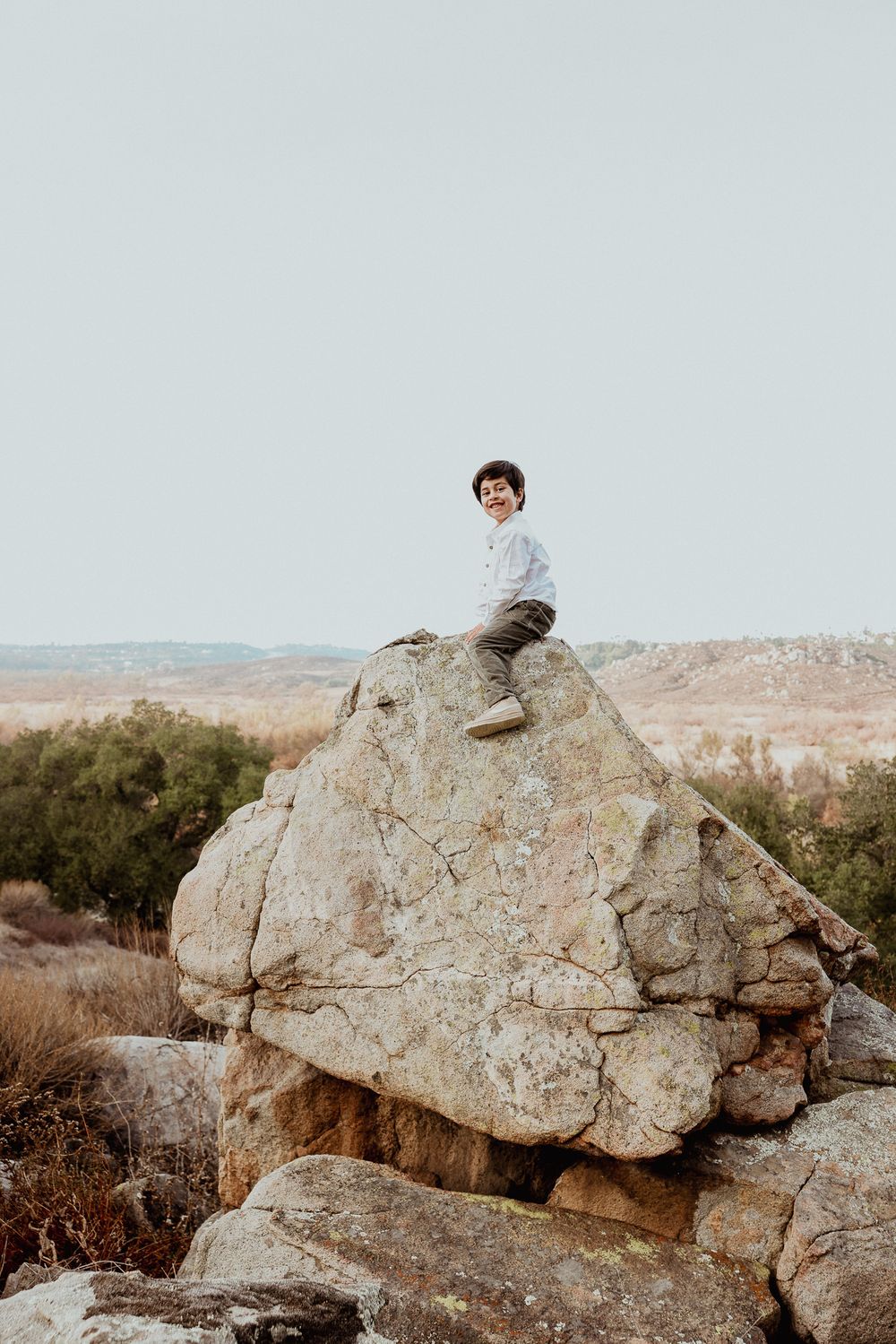 Young person sitting casually atop a large boulder in an arid landscape.