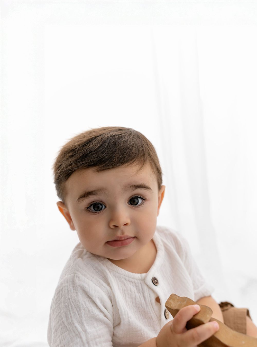 A young child in a white sweater sits against a light background while holding a teddy bear.