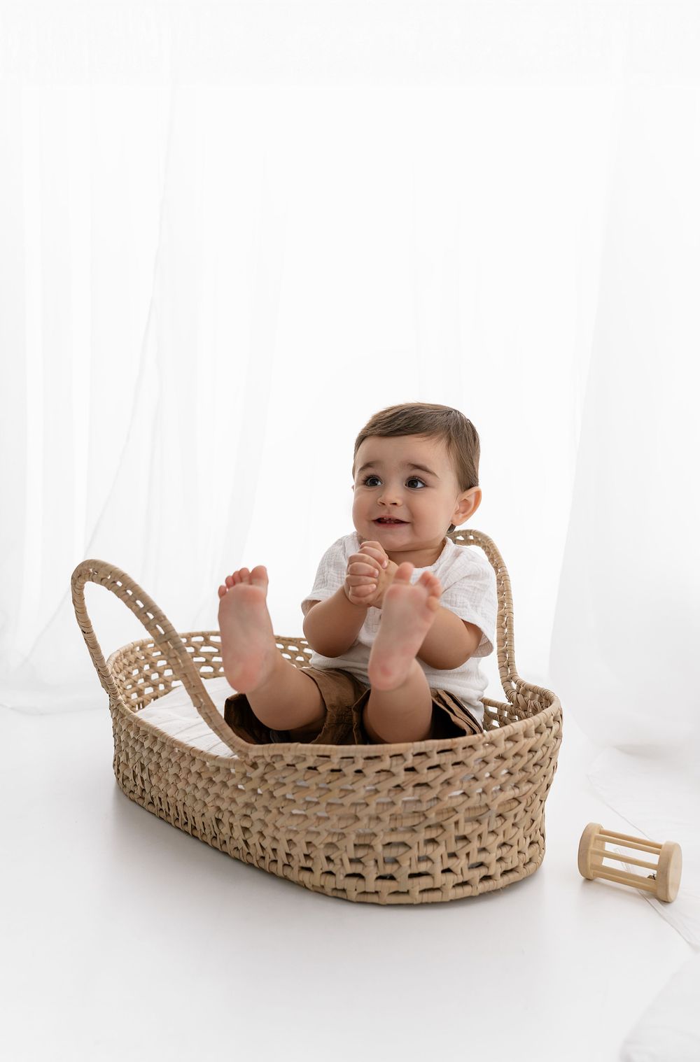 A baby sits in a woven basket on a white background wearing khaki shorts and a white top.