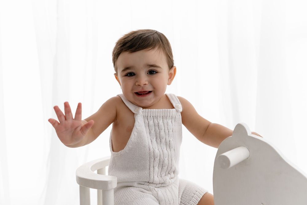 Child in white sleeveless outfit sits on white chair with hand raised playfully against bright background.