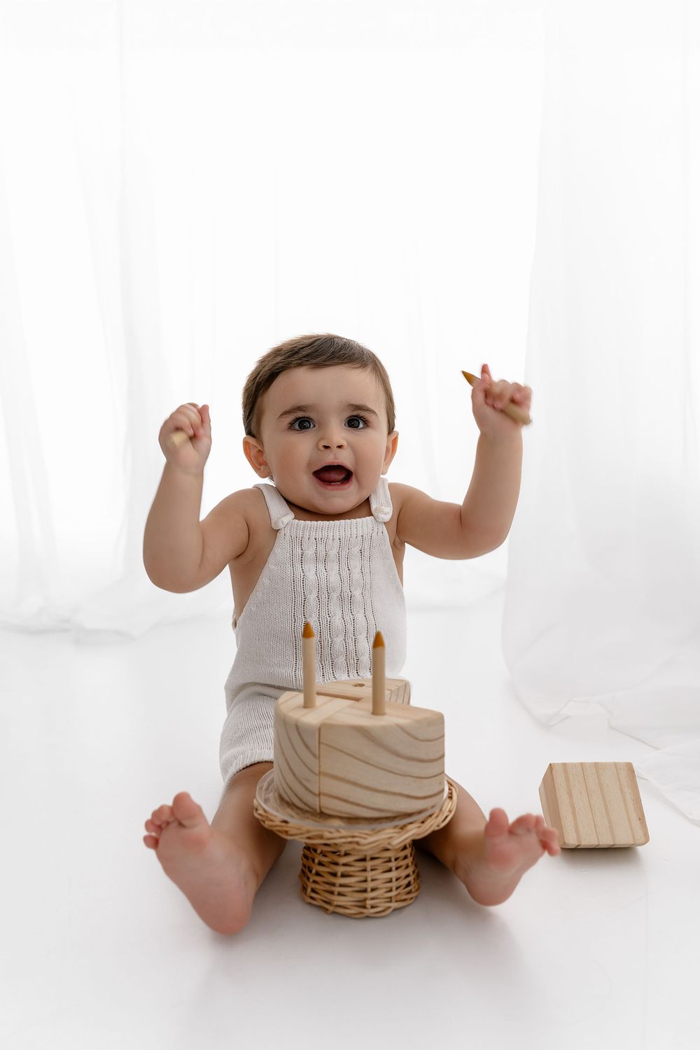 Excited baby in white romper raises arms while sitting next to wooden cake decoration on woven basket.