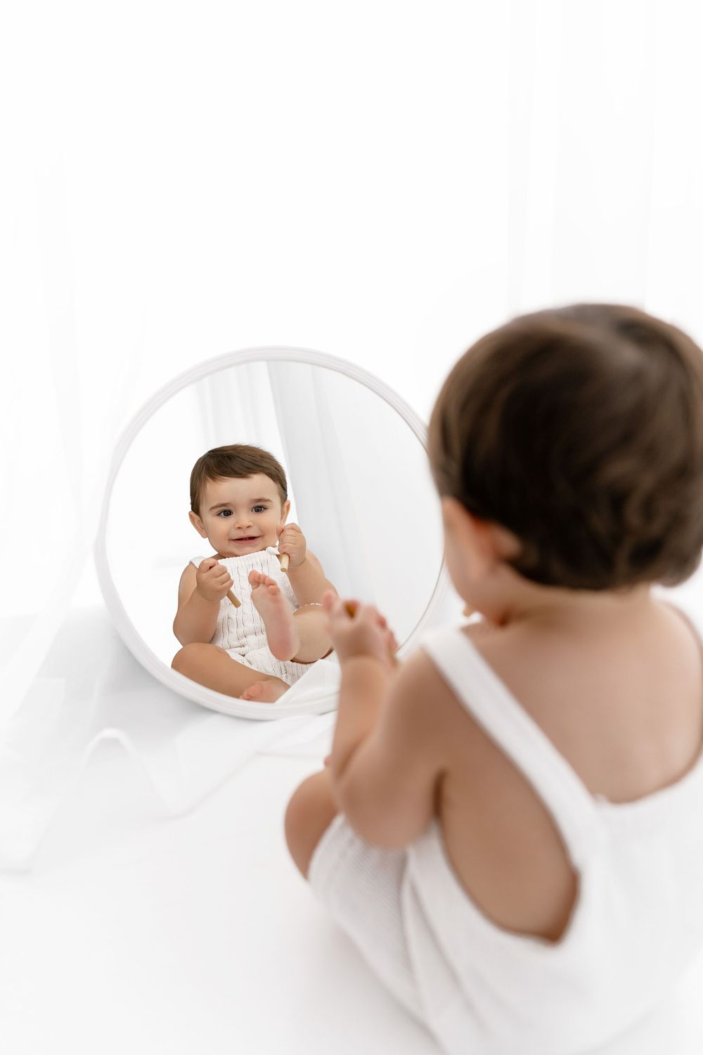 Baby in white clothing sits on floor looking at their reflection in a round mirror on white background.