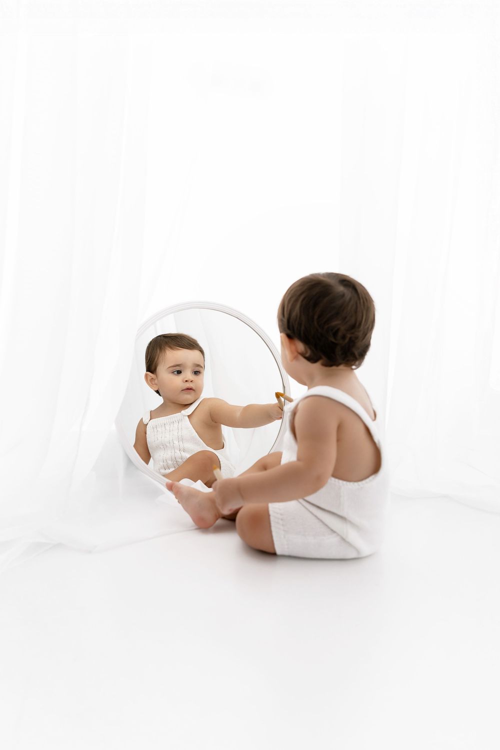 A small child in a white outfit sits in front of a mirror on a white background, reaching out to touch their reflection.