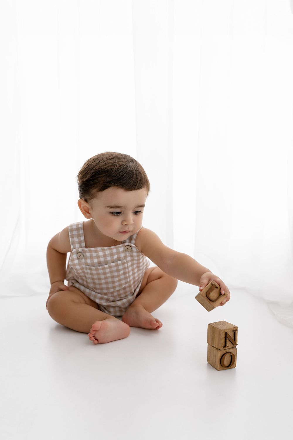 A small child in a checkered romper sits on a white floor playing with wooden alphabet blocks.