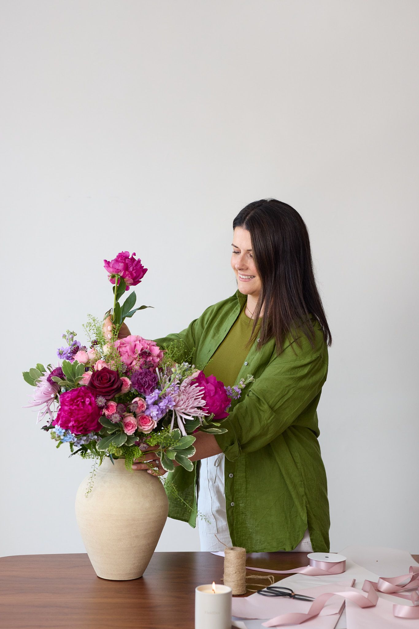 A florist in a green shirt arranges vibrant pink and purple flowers in a beige ceramic vase on a wooden table.