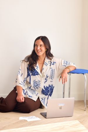 Someone in a blue and white patterned blouse sits with a laptop on the floor against a minimal white background.