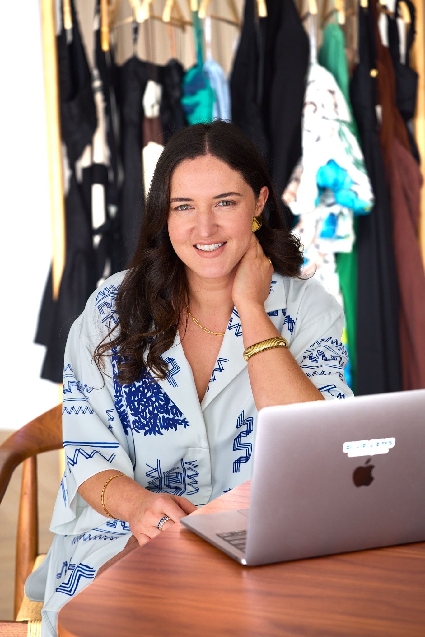 Fashion business owner working on laptop in front of clothing rack wearing blue and white patterned shirt. Personal Branding Photography.