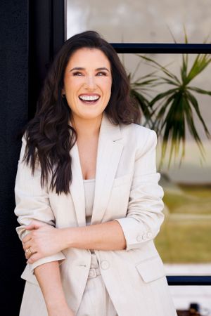 Professional portrait of someone in a cream coloured blazer smiling warmly against an modern backdrop with a plant. Personal Branding Photog