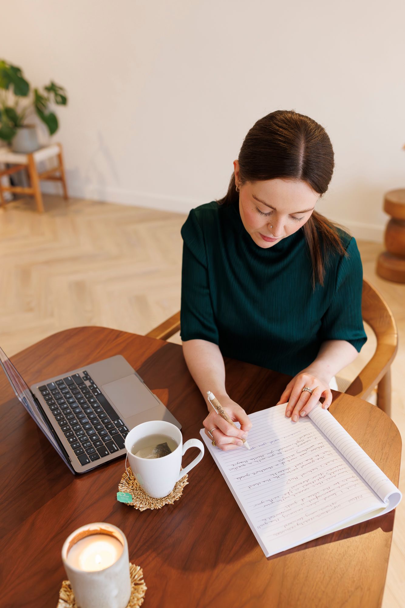 Someone in a dark green sweater writing in a notebook at a wooden table with laptop and coffee cup. Personal Branding Photography.