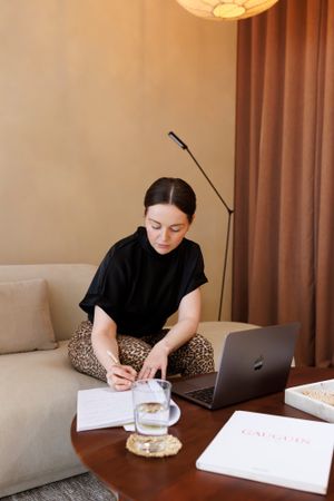 Life coach business owner in black top taking notes while working from home with laptop on couch. Personal Branding Photography.