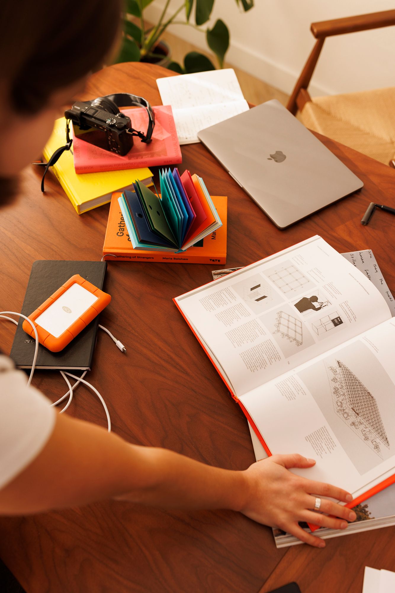 A work setup on wooden desk with MacBook, colorful notebooks, external drive and open books in warm lighting. Personal Branding Photography.
