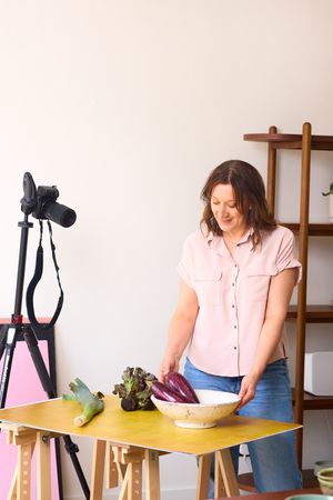 Content creator preparing food ingredients at yellow desk with camera setup and wooden coat rack in background.