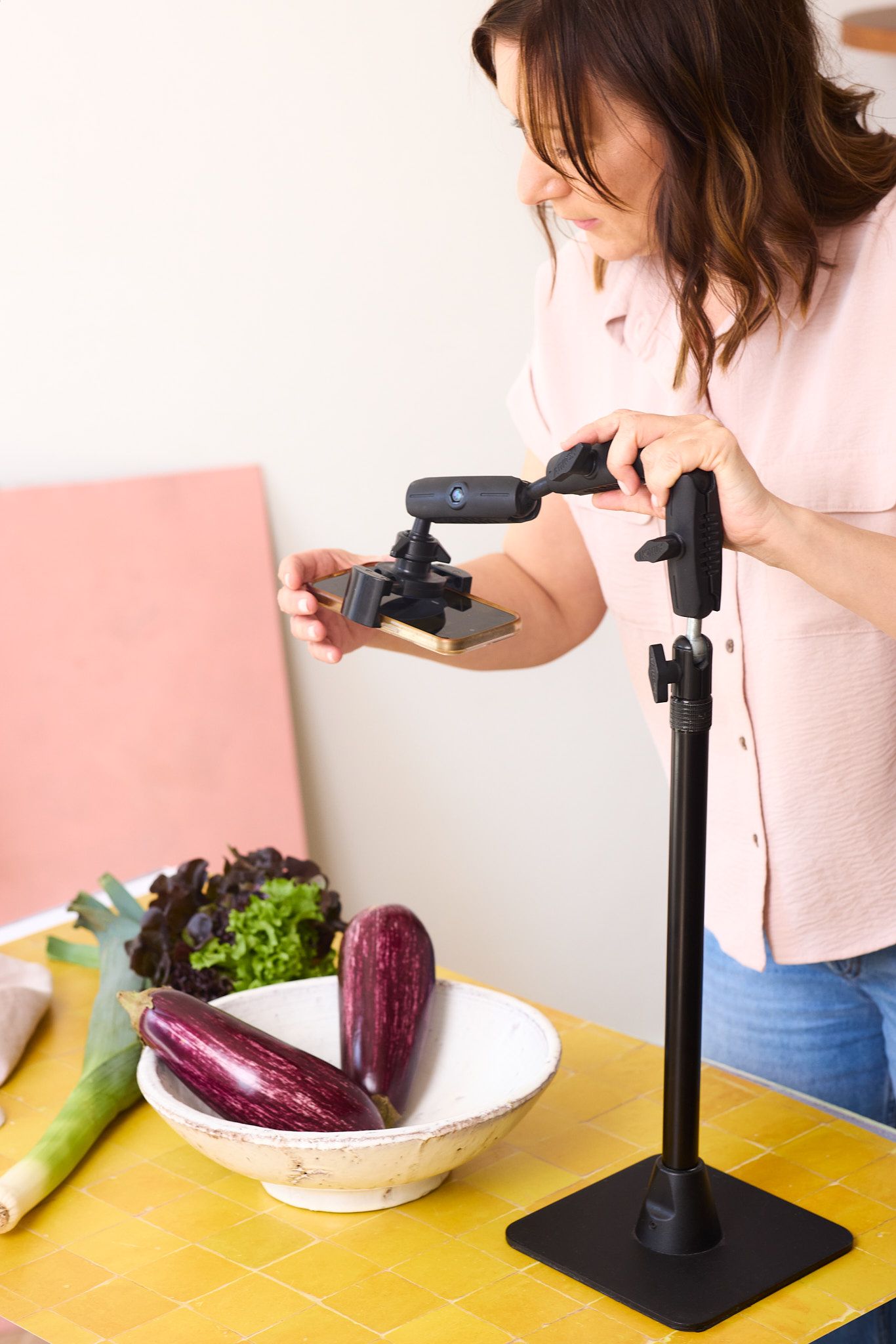 Content creator photographing fresh eggplants and produce on yellow table with smartphone mounted on adjustable stand.