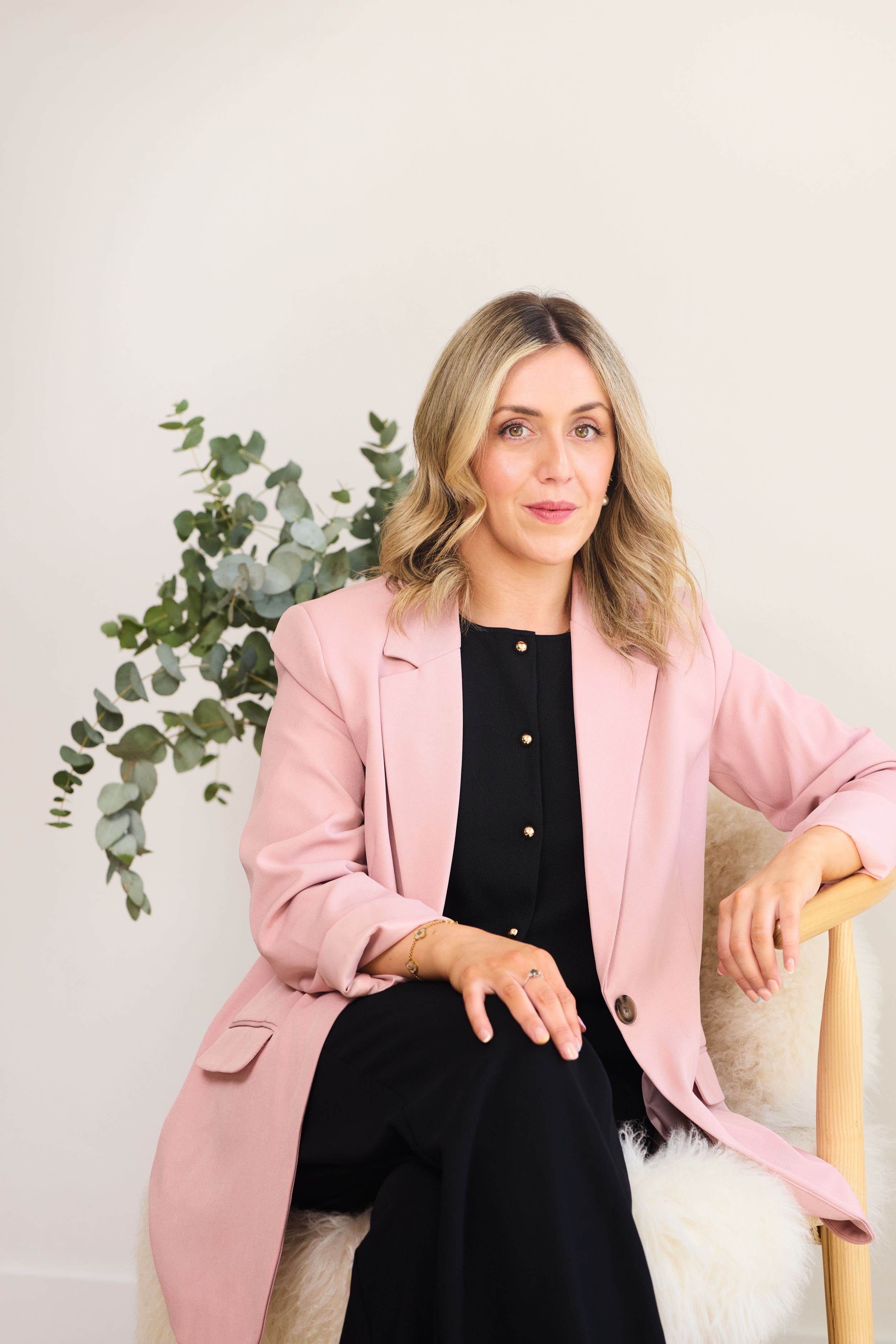 Professional portrait featuring a pink blazer over black dress with decorative eucalyptus and a cream chair in the background.