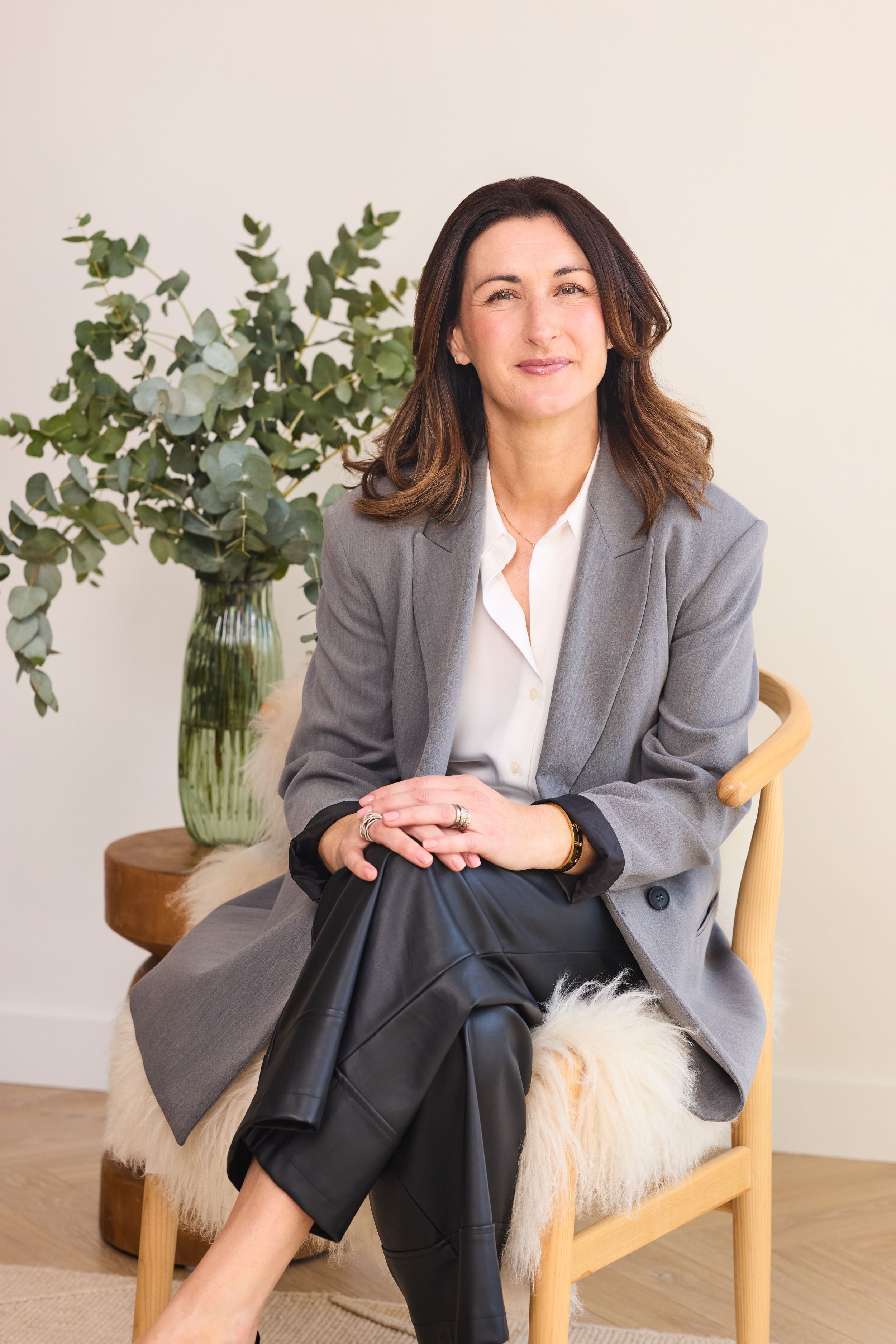 Professional wearing a gray blazer and white blouse sits in wooden chair next to eucalyptus arrangement in glass vase.