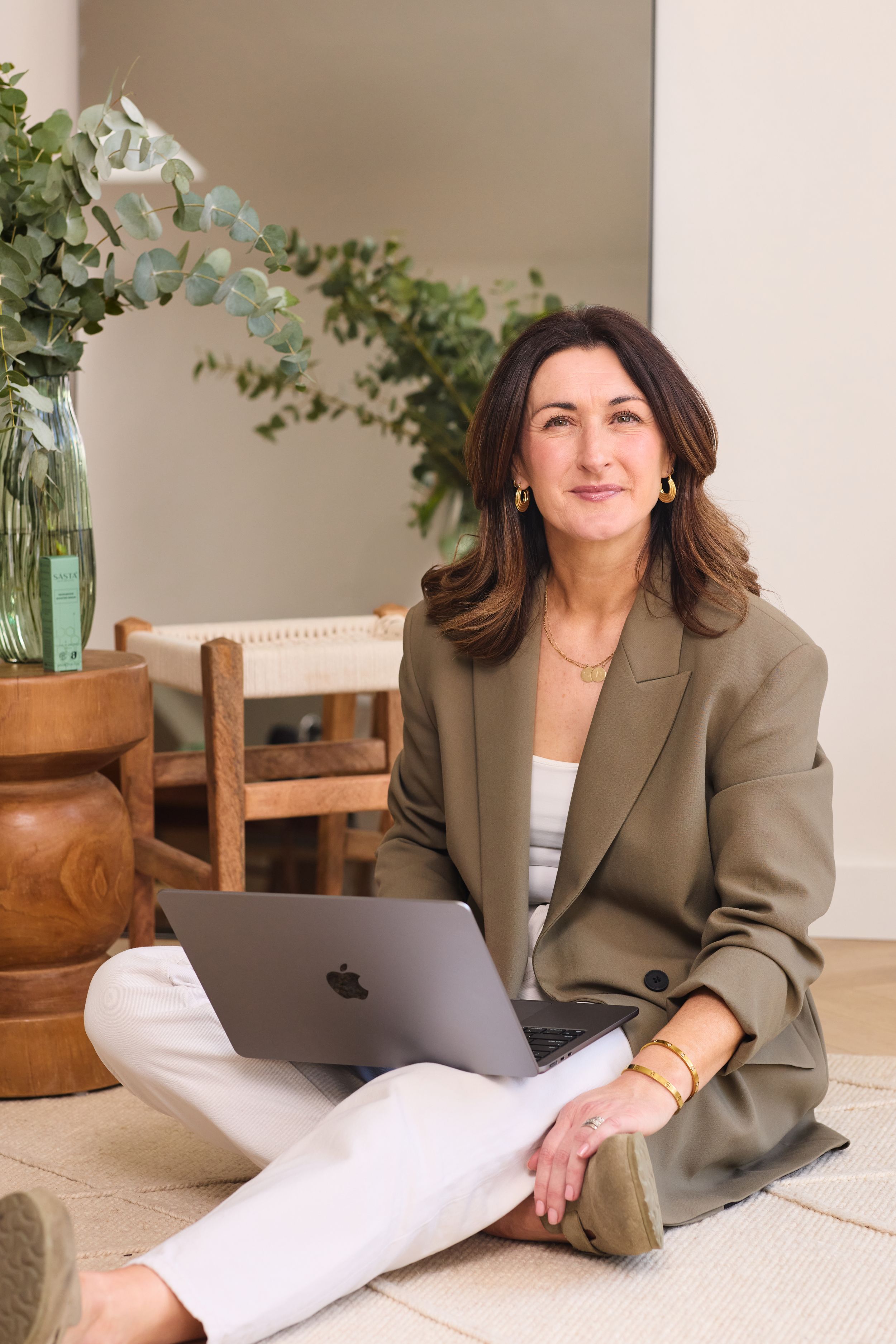 Professional seated on floor with laptop wearing olive blazer and white pants in casual home office with greenery decor.