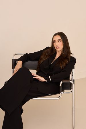 Model reclines on modern chrome chair wearing black ensemble in studio setting.