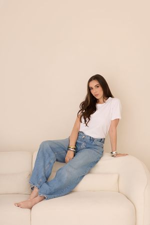 A person in a white t-shirt and blue jeans reclines on a cream colored couch in a minimalist setting.