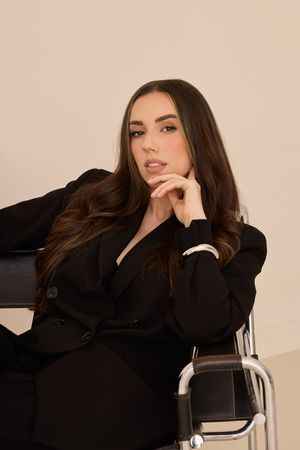 Fashion model in black blazer poses on modern chair against beige backdrop in artistic portrait series.