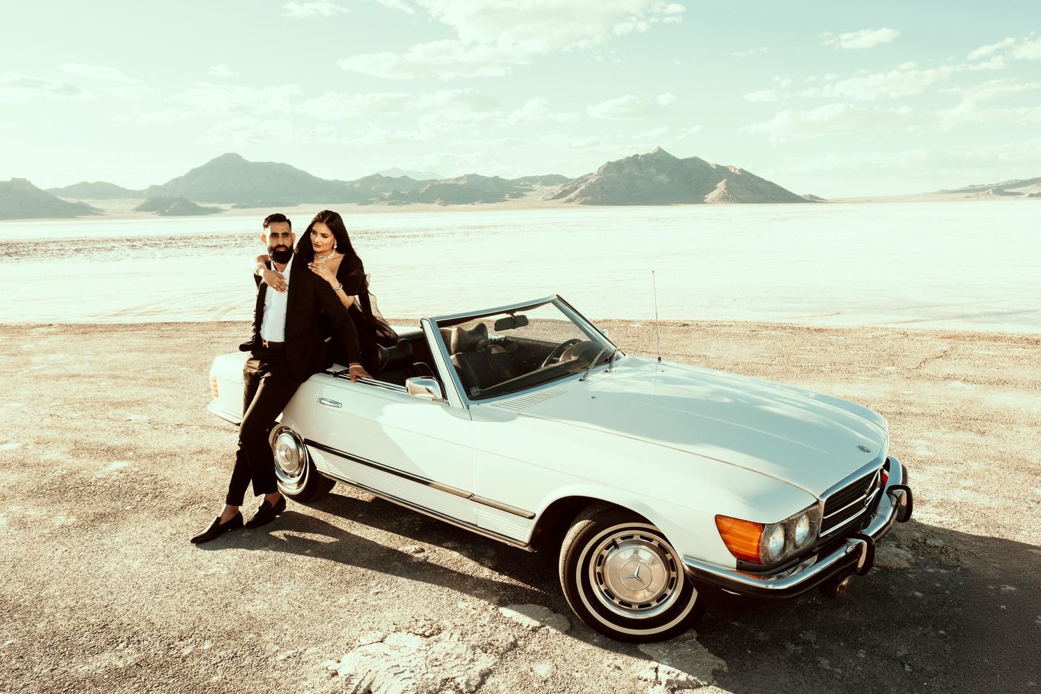 Classic silver Mercedes-Benz SL convertible parked on desert plains with mountains and cloudy sky in background.