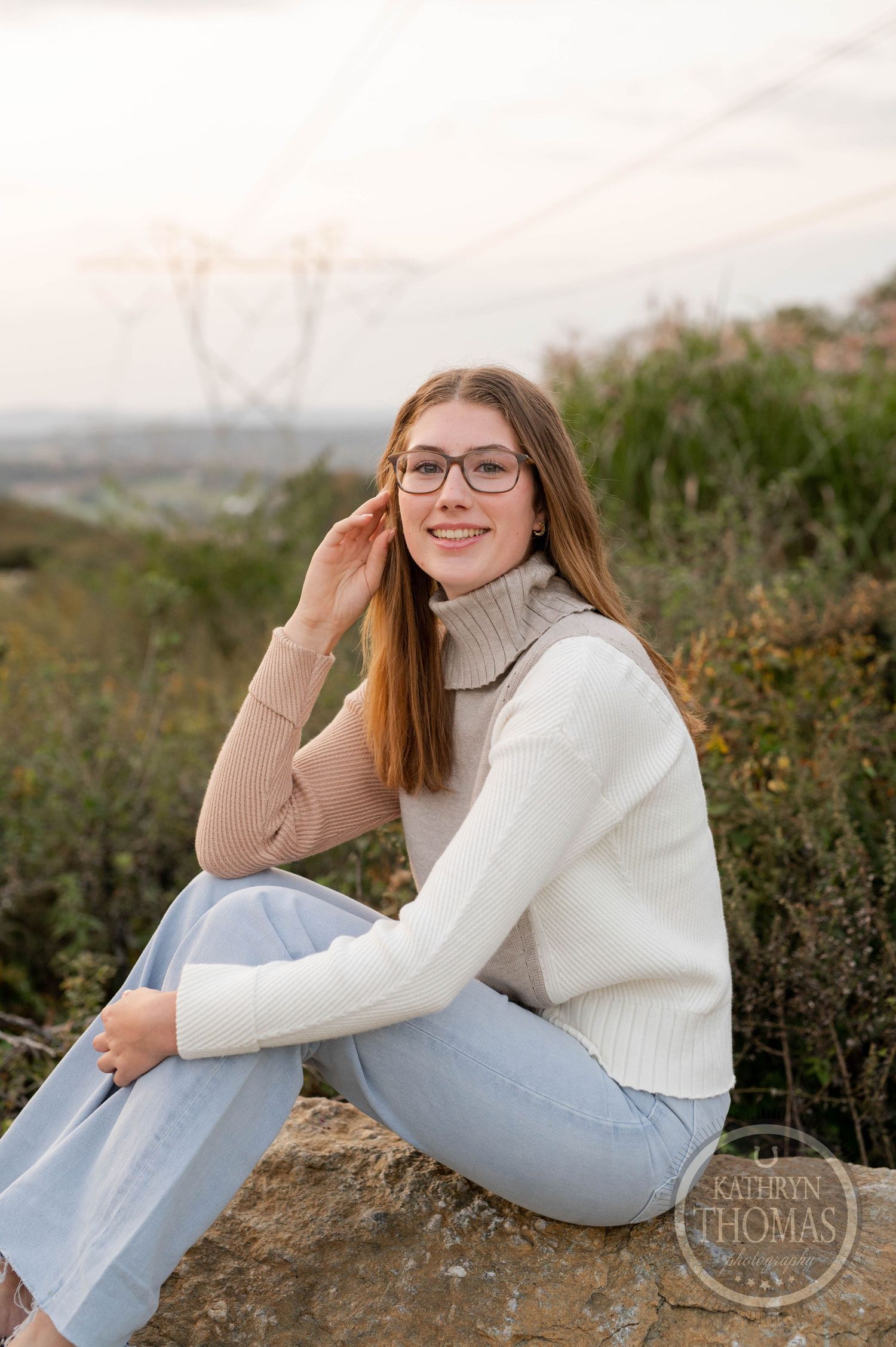 Someone in a white sweater and jeans sits on rocks with power lines visible in the background.