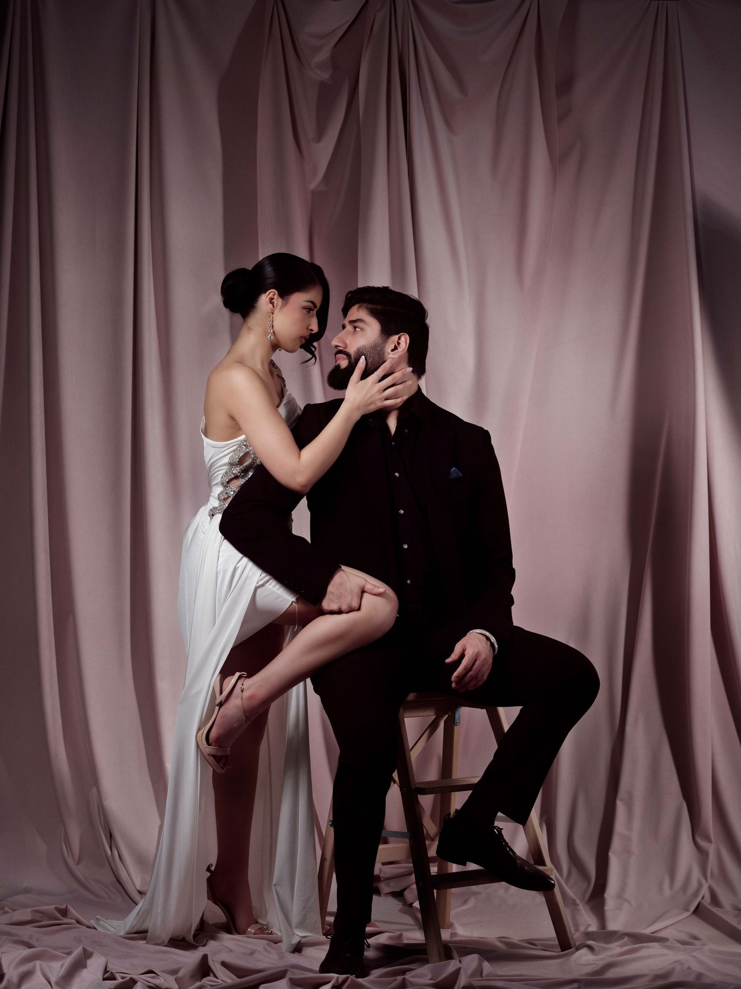 Romantic couple in dramatic pose against draped backdrop, woman in white dress and man in black suit seated on chair.