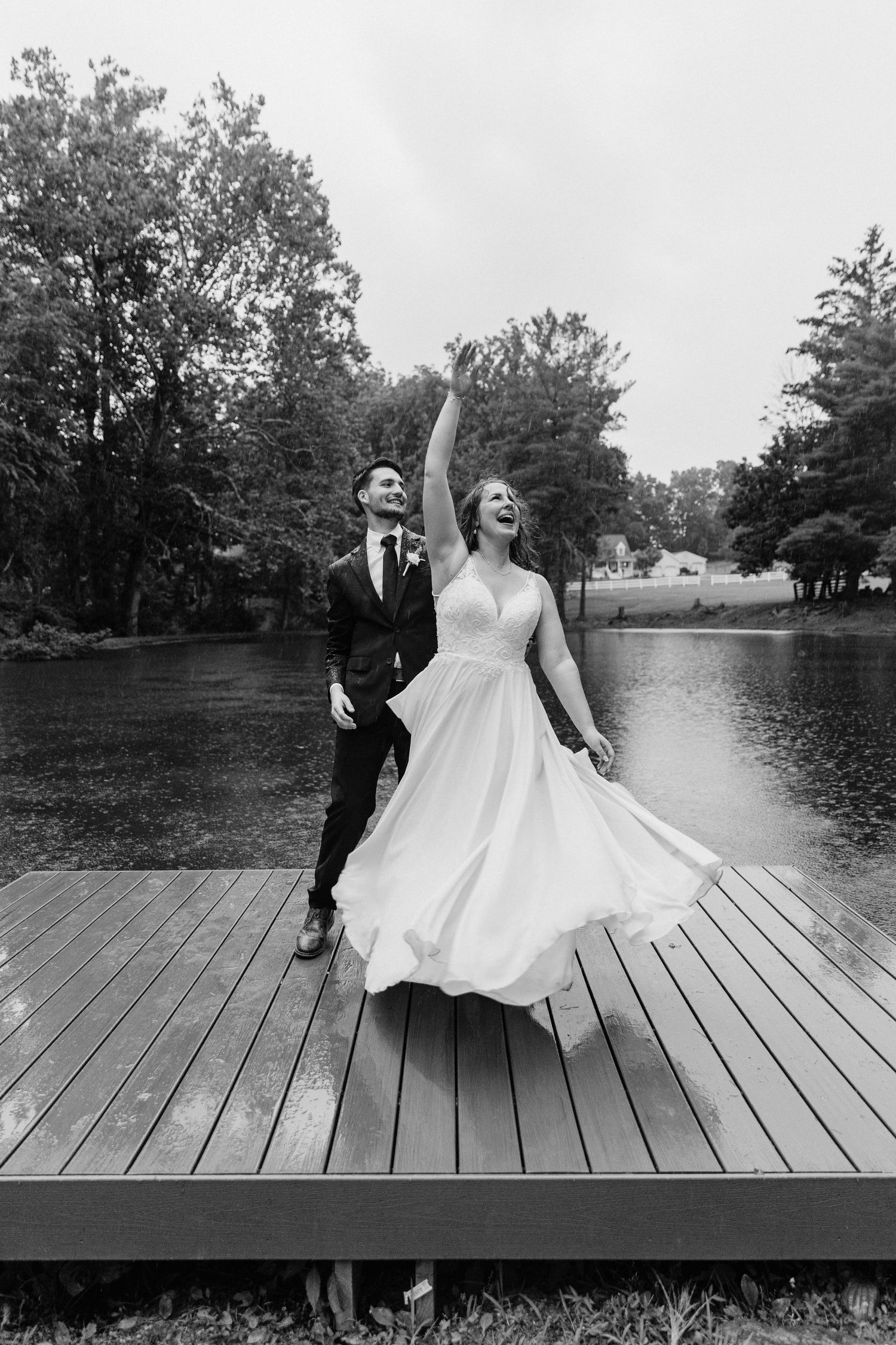 A joyful couple dances on a wooden dock during their black and white wedding photoshoot.