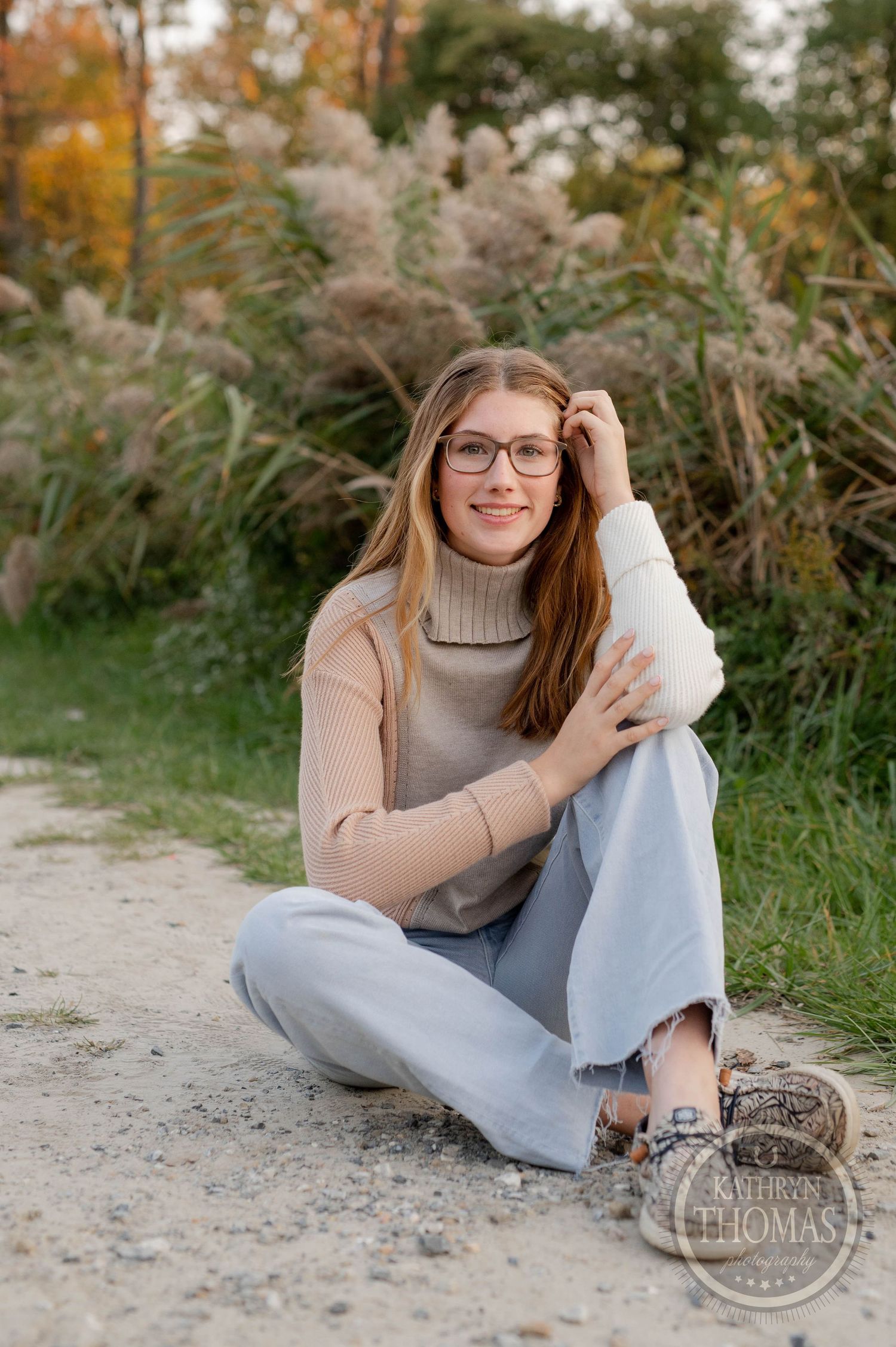 Casual sitting pose on path wearing beige sweater and glasses with autumn grasses behind.