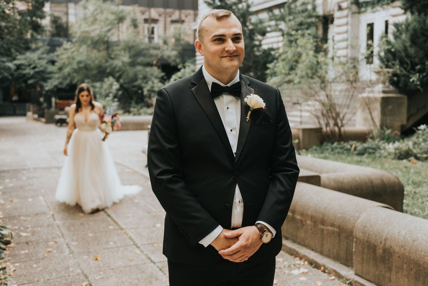 A groom in a black tuxedo and bowtie stands in the foreground while a bride in white appears in soft focus behind.