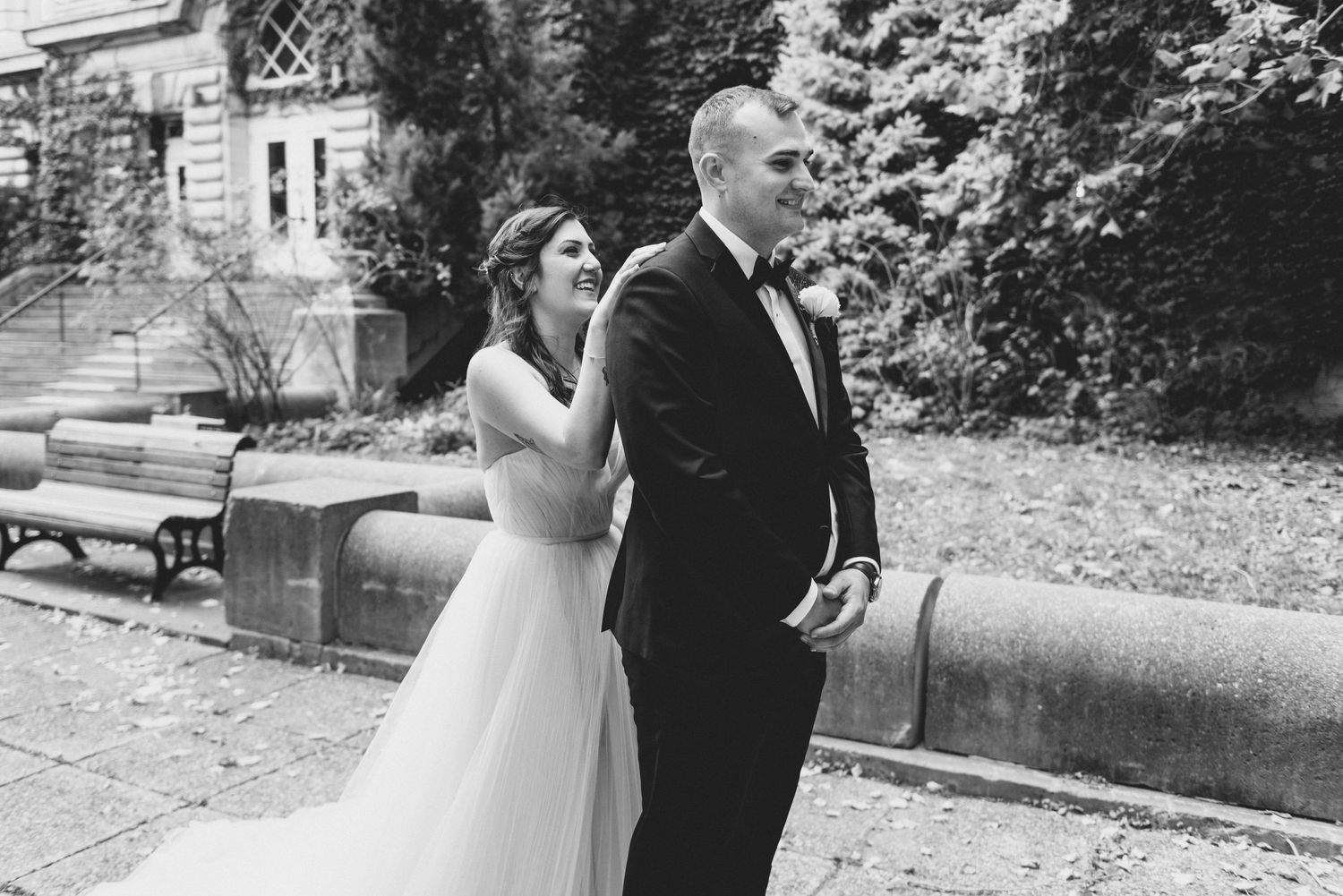 A bride in a white gown and companion in a black suit stand together outdoors in a black and white photograph.