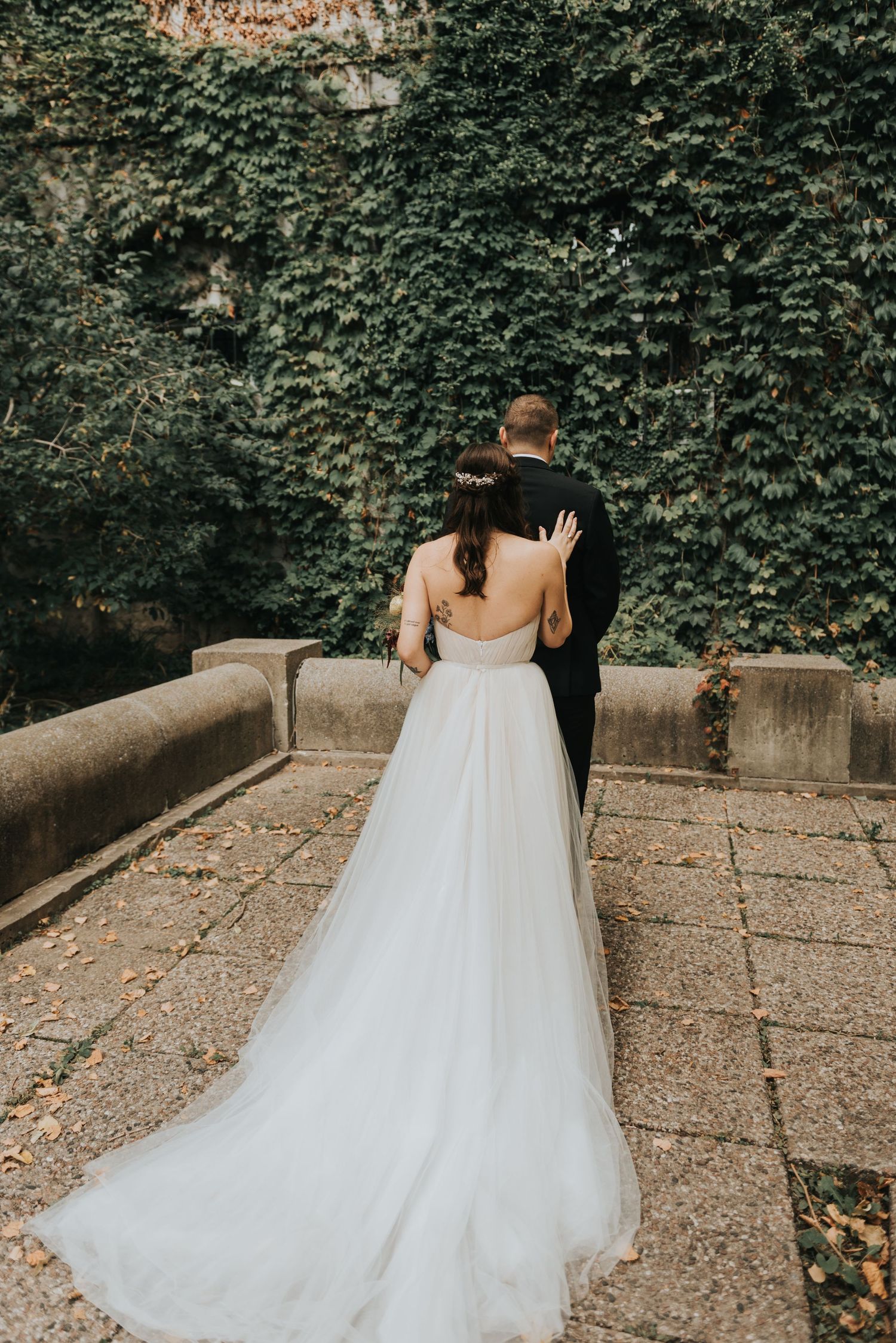 A couple in wedding attire embraces against an ivy-covered wall on a stone terrace, the bride wearing a flowing white gown.