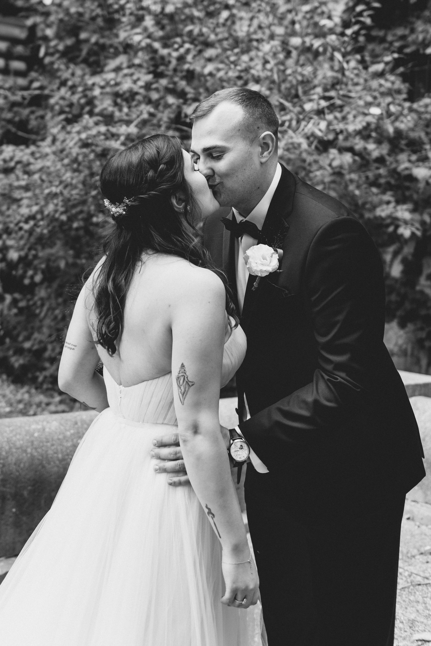 A romantic black and white wedding photo of newlyweds sharing an intimate kiss in an outdoor garden setting.