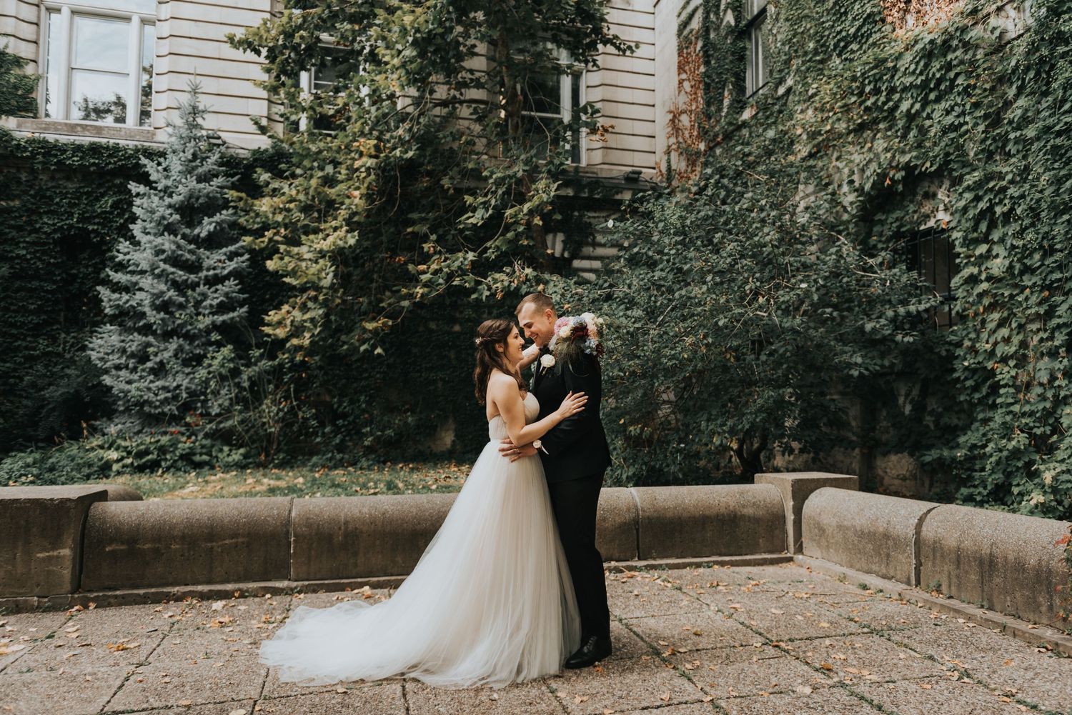A wedding couple embraces on a garden patio surrounded by lush greenery and stone walls.