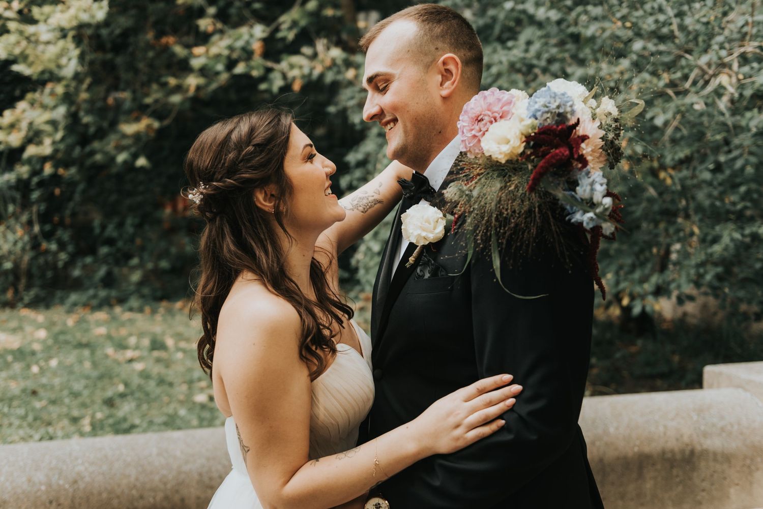 Bride and groom share an intimate moment together outdoors with floral bouquet and natural greenery in background.