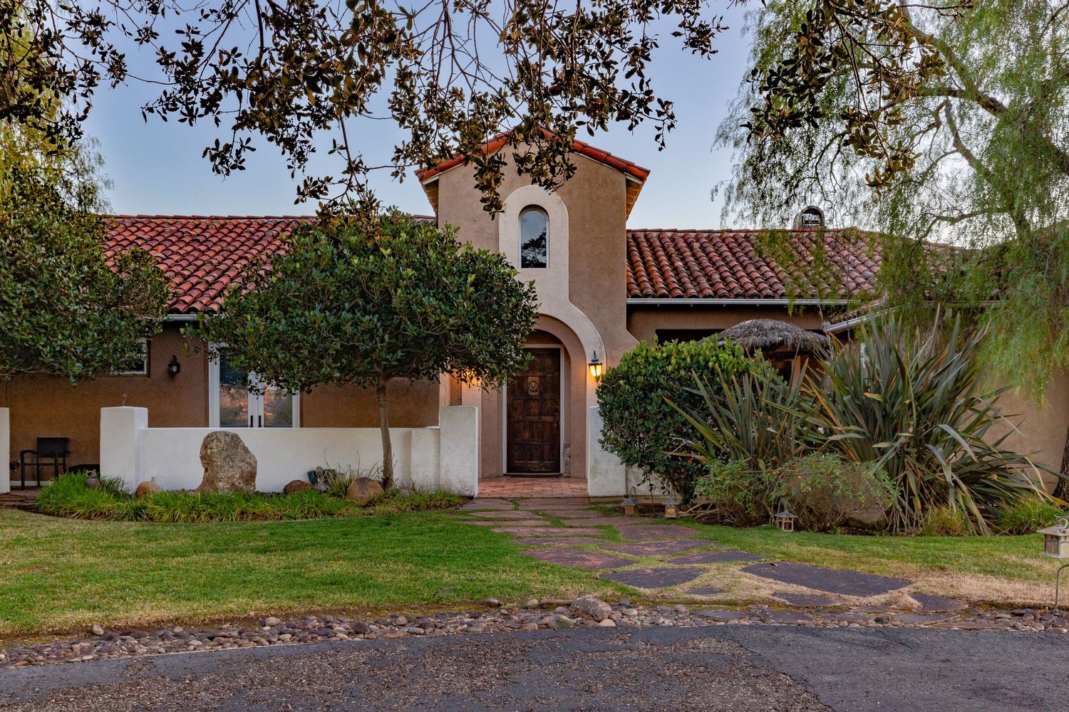 Spanish style single story home with terracotta tiled roof and arched entrance surrounded by lush landscaping at dusk.